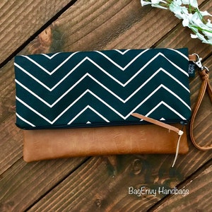 Puede incluir: Un clutch con estampado de chevron blanco y negro con una solapa de piel sintética marrón y una correa de cuero marrón. El clutch tiene cierre de cremallera y la correa está unida al lateral del clutch. El texto "BagEnvy Handbags" es visible en la esquina inferior derecha de la imagen.