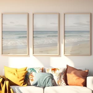 May include: Three framed beach landscape prints with light wood frames hang above a white sofa. The prints depict a serene ocean scene with a sandy beach, blue water, and a cloudy sky. Colorful throw pillows in yellow, teal, and patterned fabrics adorn the sofa.