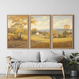 Peut inclure: Ensemble de trois peintures de paysage encadrées. Chaque œuvre représente une scène rurale avec des arbres dorés, des champs et une maison blanche sous un ciel nuageux. Elles sont au-dessus d'un canapé blanc avec un coussin gris.