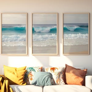 Puede incluir: Tres impresiones enmarcadas de olas del océano. Cada impresión presenta una ola turquesa rompiendo en una playa arenosa bajo un cielo nublado. Los marcos son de madera clara. Debajo, cojines coloridos sobre un sofá blanco.
