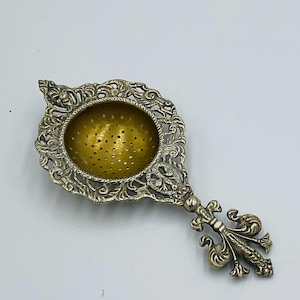 Puede incluir: Colador de té plateado ornamentado con interior dorado. El colador presenta un mango decorativo con diseño de flor de lis y filigrana intrincada alrededor del cuenco. Diseñado para té de hojas sueltas, mide aproximadamente 15 cm de largo.
