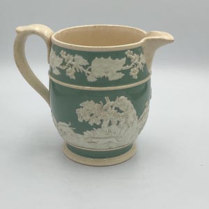 Op de afbeelding: Een groene keramische kan met witte reliëfmotieven van een wijnstok en een scène met figuren. De kan heeft een handvat en een tuit.
