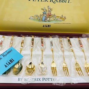 May include: A set of gold-toned spoons and forks with white ceramic handles featuring Peter Rabbit illustrations, presented in a yellow and maroon box. The set includes a blue card with the text The World of Peter Rabbit and Asahi branding.