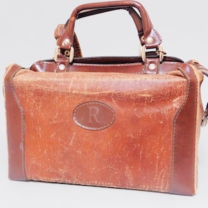 Puede incluir: Un bolso de mano vintage de cuero marrón con asa superior y forma rectangular. El bolso presenta un acabado envejecido, un pequeño emblema ovalado con la letra "R" y herrajes metálicos. El bolso tiene un diseño clásico y atemporal.