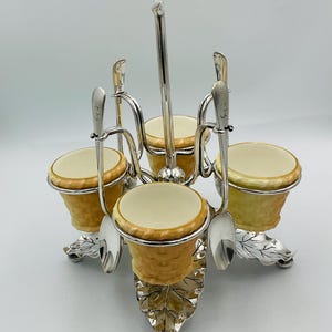 May include: A silver egg holder with four ceramic egg cups and spoons. The egg cups are decorated with a woven basket pattern in yellow and white.