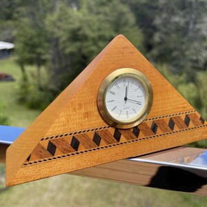 May include: A wooden pyramid-shaped clock with a gold-tone clock face. The clock has a light brown wood grain with a decorative inlaid pattern of dark brown and black diamonds along the base. The clock is set against a blurred green background.
