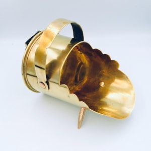 May include: A brass coal scuttle with a handle and a pointed base. The scuttle has a scalloped edge and is designed to hold coal for a fireplace.