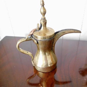 May include: A brass coffee pot with a hinged lid and a spout. The pot has a traditional Arabic design and is decorated with intricate details.