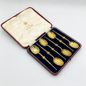 May include: A set of six gold-colored spoons in a red and blue velvet lined box. The spoons have ornate handles and detailed bowl designs. The box has a cream-colored satin lining and gold lettering.