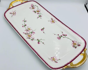 May include: A white porcelain serving tray with gold handles and a burgundy trim. The tray is decorated with a floral pattern of pink flowers and green leaves.