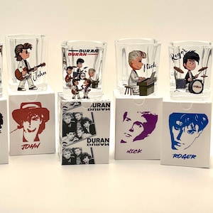 May include: Set of six clear shot glasses featuring cartoon illustrations of Duran Duran band members. Each glass has a different band member's image and name. The glasses sit on white boxes with band member portraits.