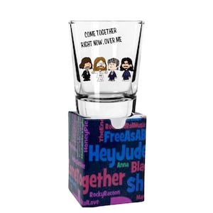 Könnte beinhalten: Ein klares Schnapsglas mit dem Text "COME TOGETHER RIGHT NOW, OVER ME" und einer Cartoon-Illustration von vier Figuren. Das Schnapsglas steht auf einer blauen Schachtel mit Songtiteln und Texten.