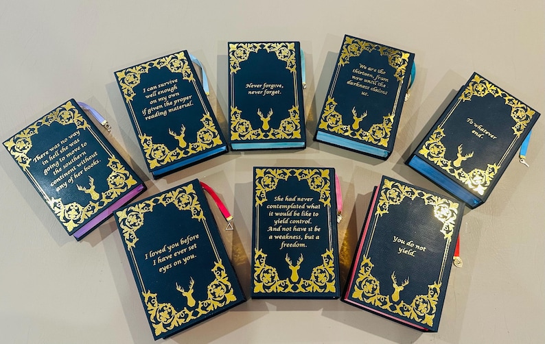 K&ouml;nnte beinhalten: Zehn kleine, dunkelblaue, gebundene B&uuml;cher mit goldenen Akzenten und goldenen Lettern. Jedes Buch hat ein anderes Zitat auf dem vorderen Umschlag. Die Zitate handeln von Liebe, Freiheit und &Uuml;berleben.