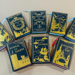 K&ouml;nnte beinhalten: Eine Sammlung von neun Hardcover-B&uuml;chern mit goldenen Schriftz&uuml;gen und Illustrationen auf dunkelblauem Hintergrund. Die Titel lauten "The Assassin's Blade", "Throne of Glass", "Crown of Midnight", "Heir of Fire", "Queen of Shadows", "Tower of Dawn", "Empire of Storms", "Kingdom of Ash".