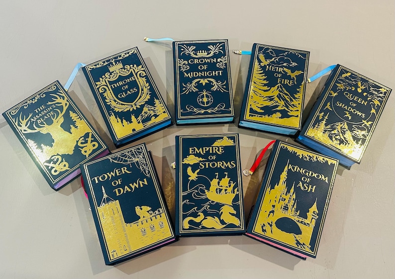 K&ouml;nnte beinhalten: Eine Sammlung von neun Hardcover-B&uuml;chern mit goldenen Schriftz&uuml;gen und Illustrationen auf dunkelblauem Hintergrund. Die Titel lauten "The Assassin's Blade", "Throne of Glass", "Crown of Midnight", "Heir of Fire", "Queen of Shadows", "Tower of Dawn", "Empire of Storms", "Kingdom of Ash".
