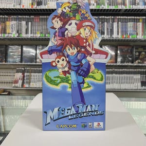 May include: A Mega Man Legends video game display. The box is blue and white, with the game title and Capcom logo. The display has a colourful illustration of the game's characters. Shelves of video games are in the background.