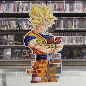 Op de afbeelding: Een kleurrijke kartonnen uitsparing met het personage Goku uit Dragon Ball Z. Goku wordt afgebeeld met stekelig geel haar, gekleed in een oranje en blauwe outfit. De uitsparing promoot "Dragon Ball Z: The Legacy of Goku II", uitgebracht in juni 2003.