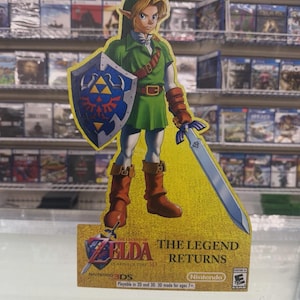 May include: A cardboard standee featuring Link from The Legend of Zelda: Ocarina of Time 3D. Link wears a green tunic, brown boots, and holds a blue Hylian shield and a sword. The base displays the game title, Nintendo 3DS logo, and text: The Legend Returns.