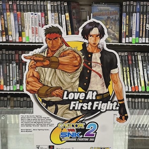K&ouml;nnte beinhalten: Werbeschild f&uuml;r das Videospiel "Capcom vs. SNK 2: Millionaire Fighting 2001" mit dem Text "Love At First Fight". Das Schild zeigt zwei Kampfspielcharaktere und wird in einem Gesch&auml;ft mit Videospielen ausgestellt.