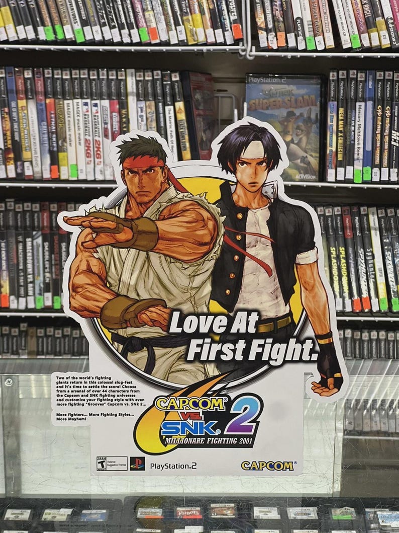 K&ouml;nnte beinhalten: Werbeschild f&uuml;r das Videospiel "Capcom vs. SNK 2: Millionaire Fighting 2001" mit dem Text "Love At First Fight". Das Schild zeigt zwei Kampfspielcharaktere und wird in einem Gesch&auml;ft mit Videospielen ausgestellt.