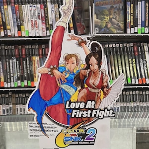 Puede incluir: Un letrero promocional del videojuego Capcom vs. SNK 2: Millionaire Fighting 2001, con dos personajes femeninos en poses de lucha. El letrero dice "Love At First Fight" e incluye los logotipos de PlayStation 2 y Capcom. El fondo muestra estanter&iacute;as de videojuegos.