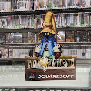 May include: A Final Fantasy IX sign featuring a character in a blue coat and a large yellow hat. The sign is in front of a display case of video games, with shelves of games in the background. The PlayStation and SquareSoft logos are also visible.