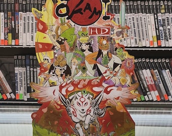 May include: A vibrant Okami video game promotional display, showcasing a white wolf with red markings and wings, alongside various characters and Japanese text. The display is positioned in front of a shelf of video games.