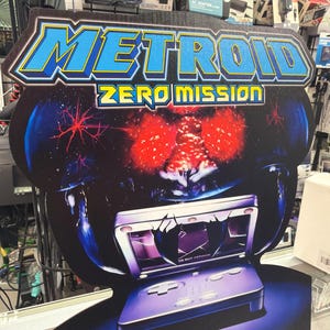 May include: A promotional cardboard display for the video game Metroid Zero Mission. The sign features the game title in blue and yellow text above a graphic of a Game Boy Advance console and a large, glowing red creature against a dark background.