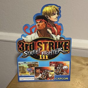 May include: A blue and yellow Street Fighter III 3rd Strike promotional display. The display features illustrations of characters Ryu and Ken, with the game title prominently displayed. The Sega Dreamcast and Capcom logos are also visible.