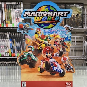 Puede incluir: Un colorido expositor de Mario Kart World con Mario y otros personajes en sus karts. El expositor tiene un borde azul y rojo con el logotipo de Nintendo Switch y una clasificación ESRB.