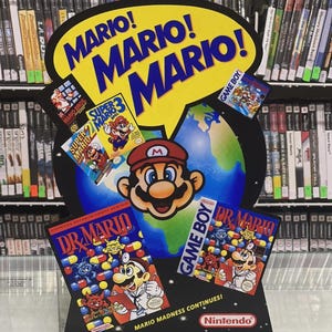 Può includere: Un colorato espositore di Mario con il testo "MARIO! MARIO! MARIO!" in un fumetto giallo. L'espositore include immagini di cartucce di videogiochi di Mario, il personaggio di Mario e il logo Nintendo. Lo sfondo mostra scaffali di videogiochi.