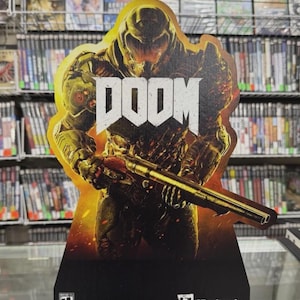 Puede incluir: Un recorte de cartón del videojuego "DOOM", que representa a un personaje con una escopeta sobre un fondo de fuego. El logotipo del juego, "DOOM", está en letras blancas. El recorte tiene una base negra y el logotipo de "Bethesda".