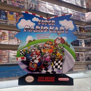 May include: A Super Mario Kart promotional sign featuring Mario, Luigi, Princess Peach, Bowser, Donkey Kong, and Yoshi racing on a track. The text reads Super Mario Kart, Where racing becomes an adventure!, Super Nintendo Entertainment System, and Nintendo.