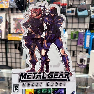 May include: A die-cut cardboard display for the Game Boy Color game Metal Gear Ghost Babel. It features illustrations of two characters in tactical gear, the game title, an ESRB E rating, Konami logo, and three small screenshots of gameplay.