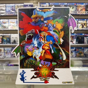 May include: A vertical display stand featuring the Vampire Savior: The Lord of Vampire game artwork. The colorful illustration depicts various anime-style characters, including a central figure in a red hood, with the game title and Capcom logo below.