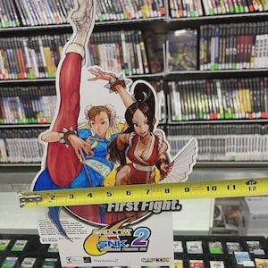 Puede incluir: Un expositor de cart&oacute;n con dos personajes femeninos del videojuego Capcom vs. SNK 2. El expositor mide aproximadamente 25 cm de alto y muestra el logotipo y el t&iacute;tulo del juego, "First Fight". El fondo incluye estanter&iacute;as de carcasas de videojuegos.