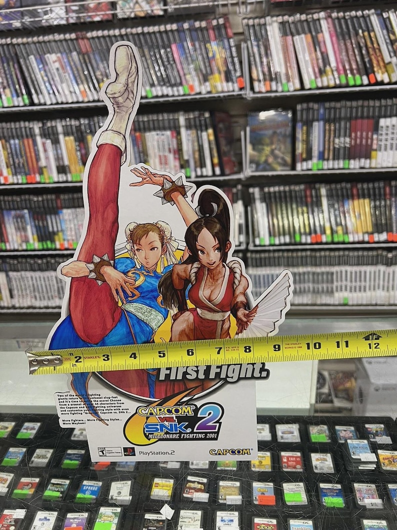 Puede incluir: Un expositor de cart&oacute;n con dos personajes femeninos del videojuego Capcom vs. SNK 2. El expositor mide aproximadamente 25 cm de alto y muestra el logotipo y el t&iacute;tulo del juego, "First Fight". El fondo incluye estanter&iacute;as de carcasas de videojuegos.
