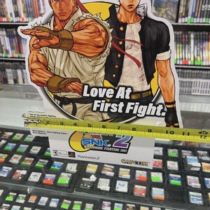 K&ouml;nnte beinhalten: Ein Werbedisplay f&uuml;r das Videospiel SNK vs. Capcom: SVC Chaos, mit Illustrationen von zwei Kampfspielcharakteren. Das Display ist etwa 25 cm breit und enth&auml;lt den Text "Love At First Fight."