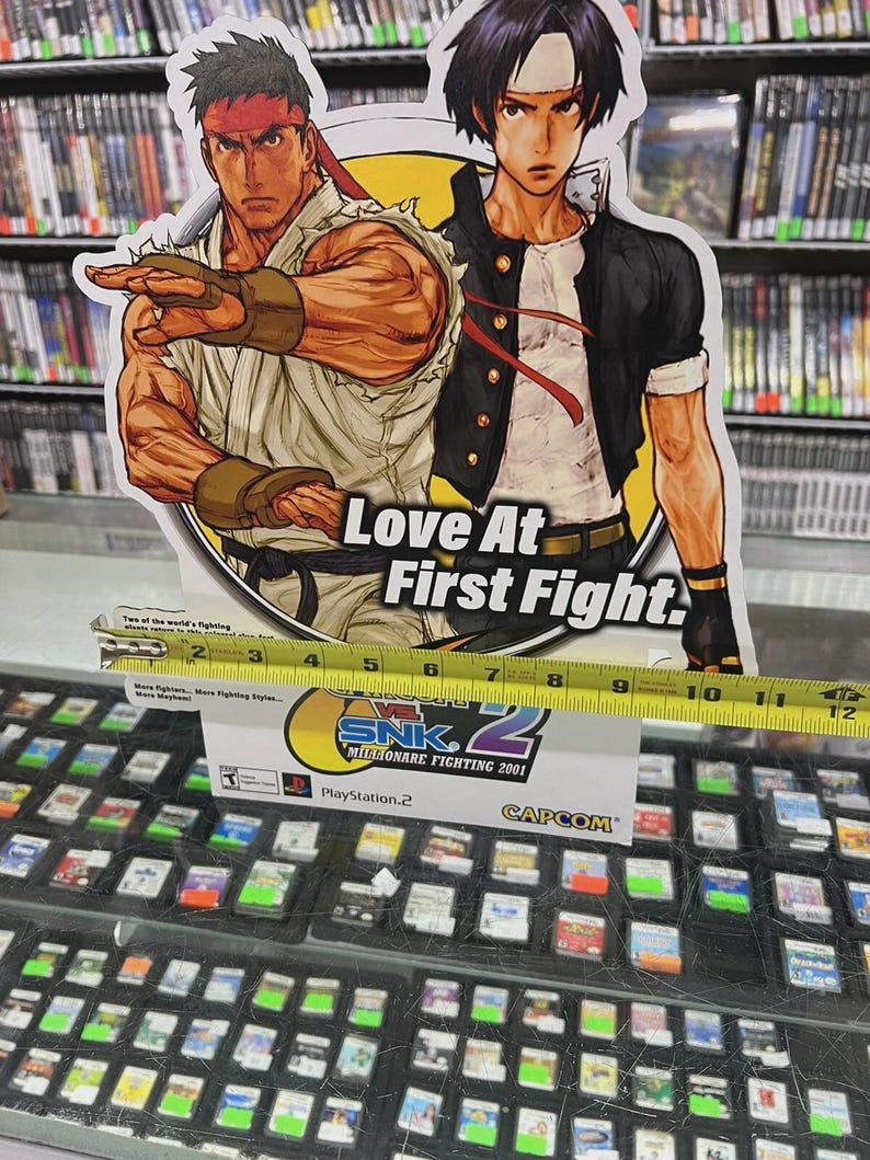 K&ouml;nnte beinhalten: Ein Werbedisplay f&uuml;r das Videospiel SNK vs. Capcom: SVC Chaos, mit Illustrationen von zwei Kampfspielcharakteren. Das Display ist etwa 25 cm breit und enth&auml;lt den Text "Love At First Fight."