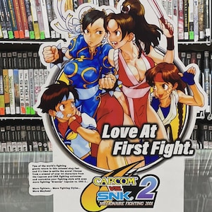 Könnte beinhalten: Ein Werbeplakat für das Videospiel "Capcom vs. SNK 2: Millionaire Fighting 2001" mit vier Charakteren im Anime-Stil. Das Plakat trägt den Text "Love At First Fight" und das PlayStation 2-Logo.