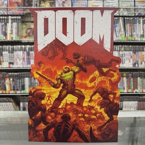 May include: A "DOOM" video game promotional sign with a red backdrop and the game's white logo. The artwork shows a battle scene with a green and brown armored character fighting demons in a fiery setting. Shelves of video games are visible in the background.