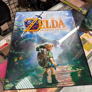 May include: A framed poster featuring The Legend of Zelda: Ocarina of Time artwork. The image displays a character with a green tunic and cap, carrying a shield and blue instrument, standing on a forest path. The title text is printed in gold.
