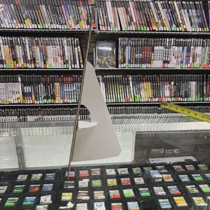 K&ouml;nnte beinhalten: Eine Ladenauslage mit Videospielen und Spielkassetten. Regale sind mit Spielh&uuml;llen gef&uuml;llt, und eine Glasvitrine darunter enth&auml;lt zahlreiche Spielkassetten. Ein Pappschild steht vor der Auslage.