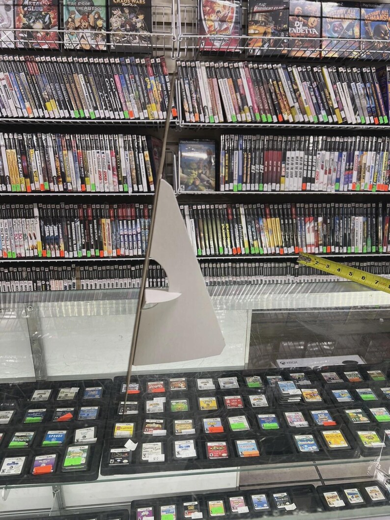 K&ouml;nnte beinhalten: Eine Ladenauslage mit Videospielen und Spielkassetten. Regale sind mit Spielh&uuml;llen gef&uuml;llt, und eine Glasvitrine darunter enth&auml;lt zahlreiche Spielkassetten. Ein Pappschild steht vor der Auslage.