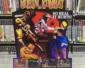 May include: A vintage "Mortal Kombat" arcade game display featuring the game's logo and characters. The display includes the text "SO REAL IT HURTS!" and "MIDWAY". The background shows shelves of video game cases.