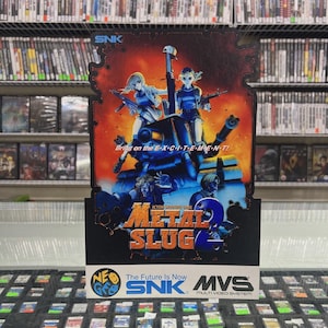 May include: Metal Slug 2 arcade game box art. The artwork features two female characters holding guns, a blue tank, and the game's title in gold. The SNK logo is at the top, with the Neo Geo MVS logo at the bottom.
