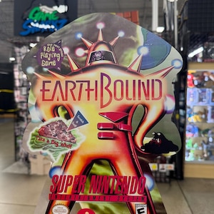May include: A promotional cardboard standee for the Super Nintendo Entertainment System game EarthBound. The display features a central orange alien figure, the game title, the text 'the next great Role Playing Game', and a 'Scratch & Sniff' sticker.