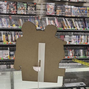 K&ouml;nnte beinhalten: Ein Pappaufsteller mit zwei Figuren steht vor Regalen voller Videospielh&uuml;llen. Die H&uuml;llen sind &uuml;berwiegend schwarz mit farbenfrohen Spielgrafiken. Darunter befindet sich eine Vitrine mit kleineren Spielkassetten. Ein Ma&szlig;band ist zu sehen.