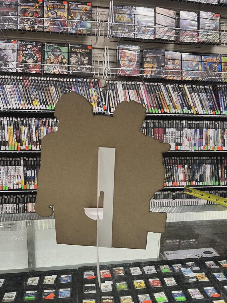 K&ouml;nnte beinhalten: Ein Pappaufsteller mit zwei Figuren steht vor Regalen voller Videospielh&uuml;llen. Die H&uuml;llen sind &uuml;berwiegend schwarz mit farbenfrohen Spielgrafiken. Darunter befindet sich eine Vitrine mit kleineren Spielkassetten. Ein Ma&szlig;band ist zu sehen.