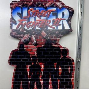 May include: A Super Street Fighter II sign featuring the Capcom logo and game title in red and blue. The background is a brick wall with silhouettes of characters. Includes Nintendo and Sega Genesis logos.